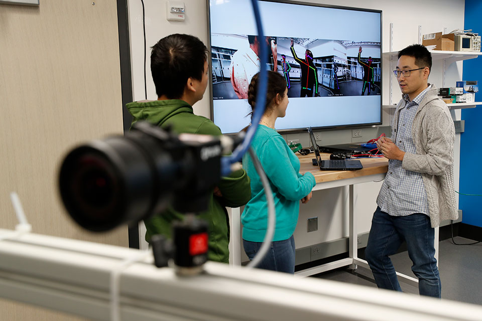 Three people talk in front of a large monitor, behind a closeup of a 3D capture camera rig