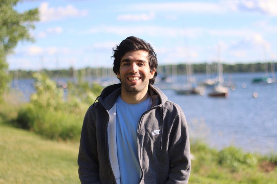 Burhaneddin Yaman wearing a grey zip-up hoodie over a light T-shirt, standing in a grassy area in front of a lake, under a blue sky 