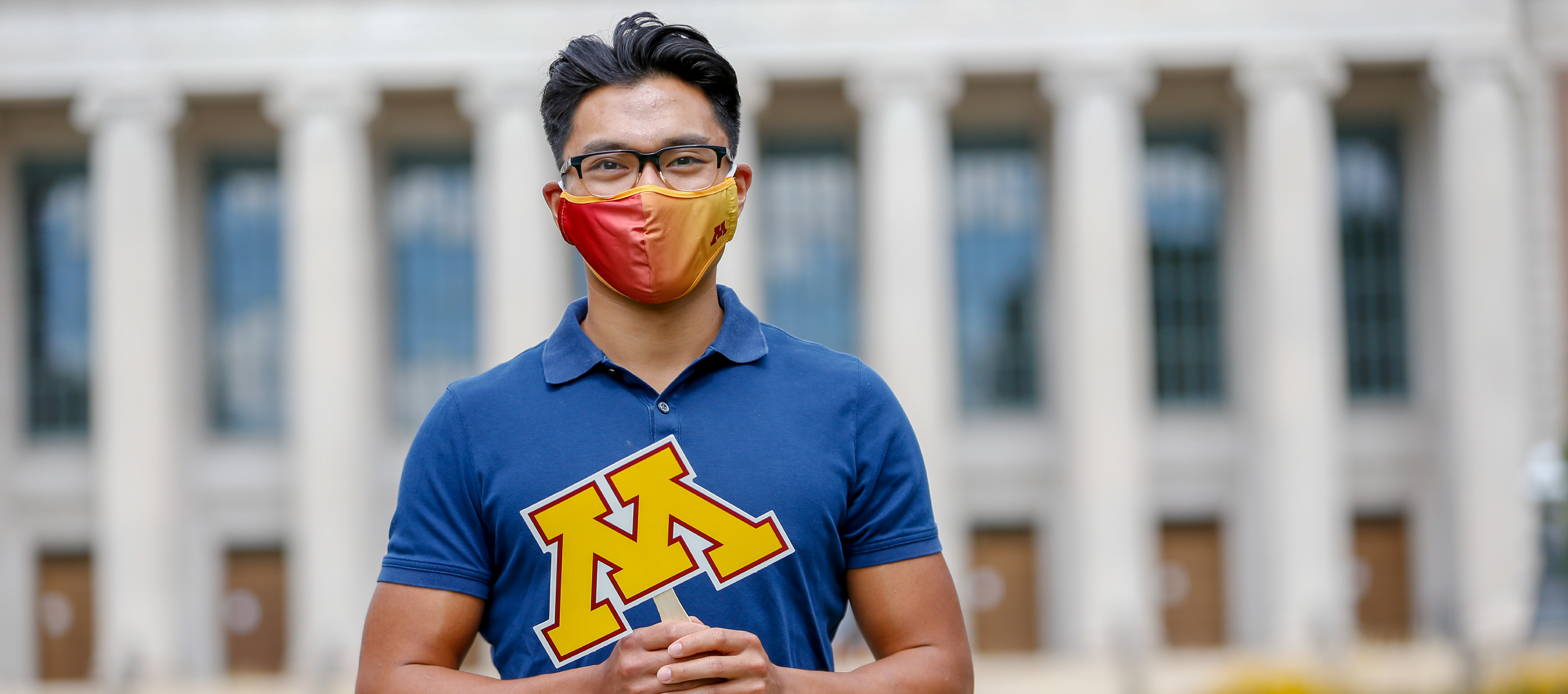 Photo of a student wearing a U of M branded face mask and holding the U of M's signature "M" logo on a stick. Student is outside with a neoclassical academic building in the background.