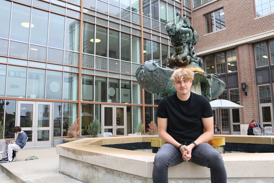 Chase Anderson in a black shirt and blue jeans sitting outdoors with his back to water fountain with a copper patina