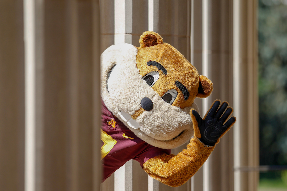 Goldy Gopher waving from behind a building column.