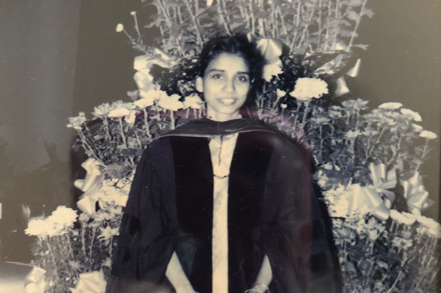 Professor Pushpavati Paladugu in her graduation robes standing in front of a floral arrangement