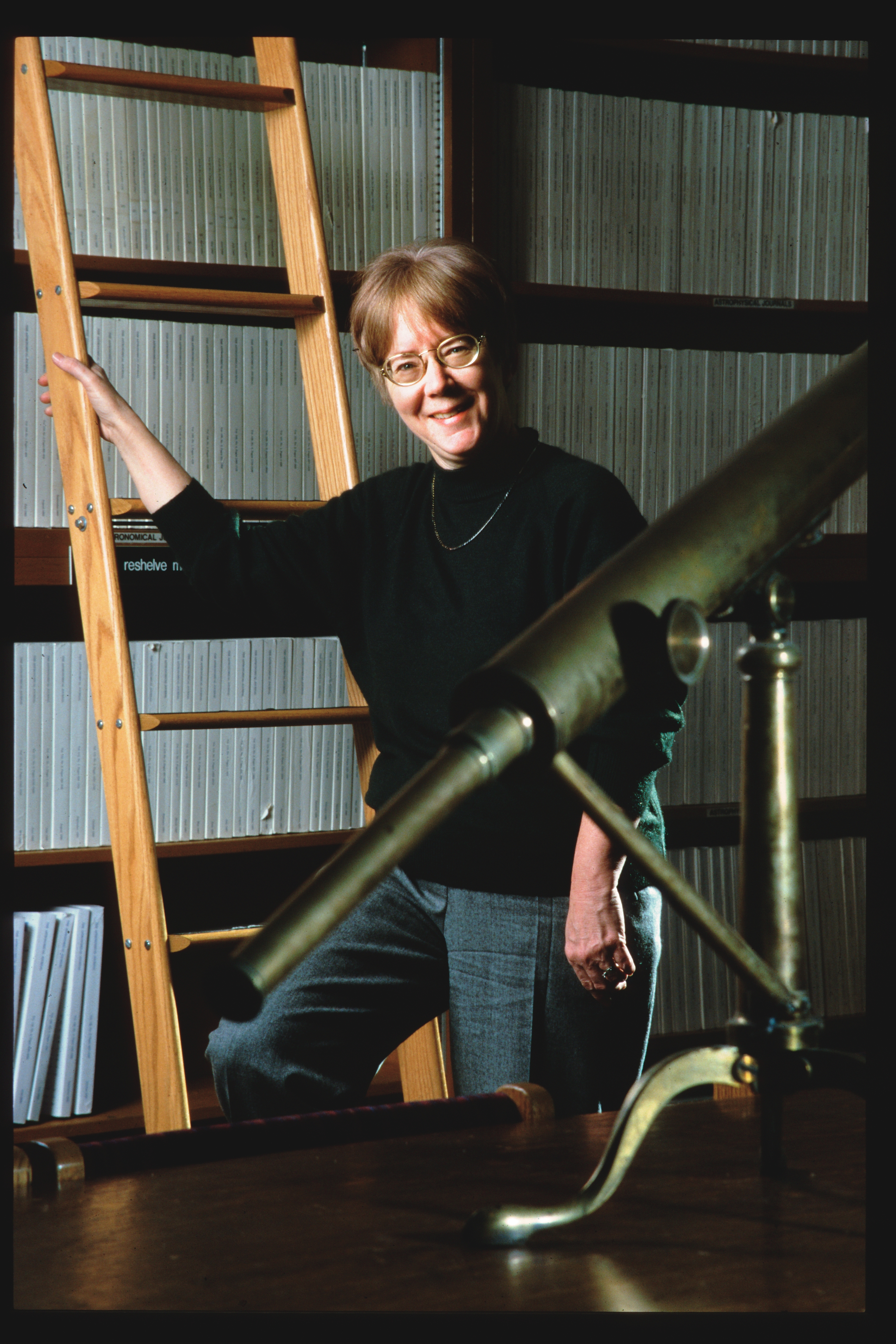 Smiling woman standing in front of a library holding onto a ladder. There is a telescope in the foreground.