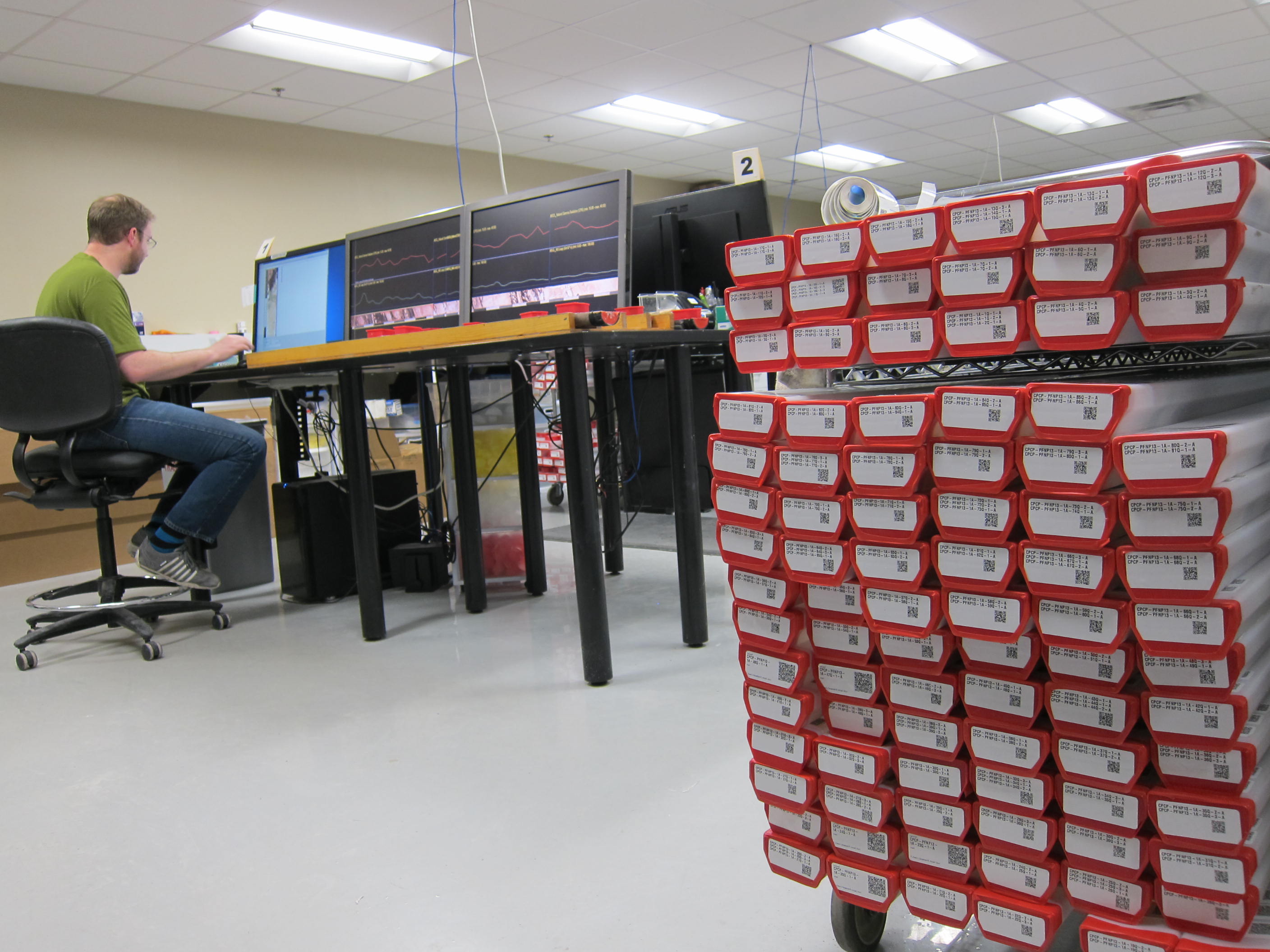 Cart full of D tubes with red caps indicating they are archive halves of the cores in the foreground with a researcher in the background analyzing data on a computer