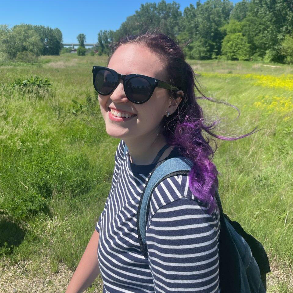 A white woman with purple hair smiles in a sunny field