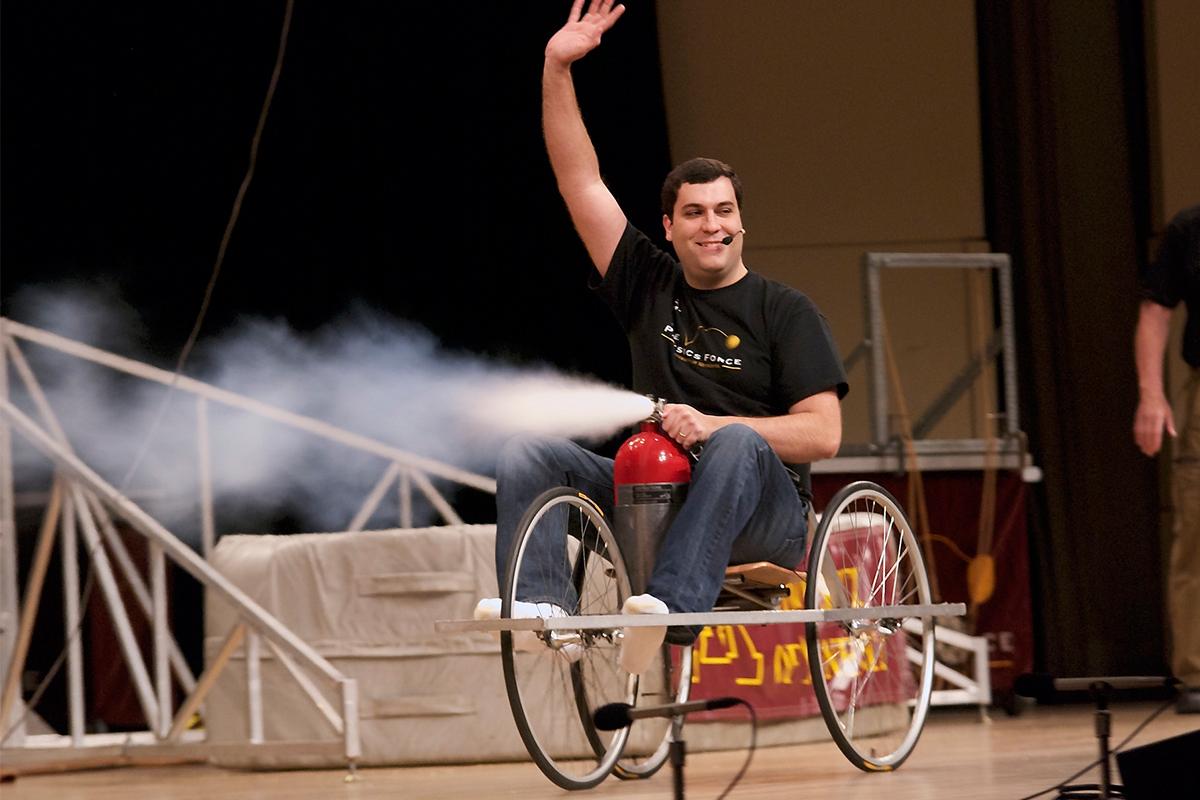 Man smiling and waving riding on a home made rocket car made from a fire extinguisher and a go cart.