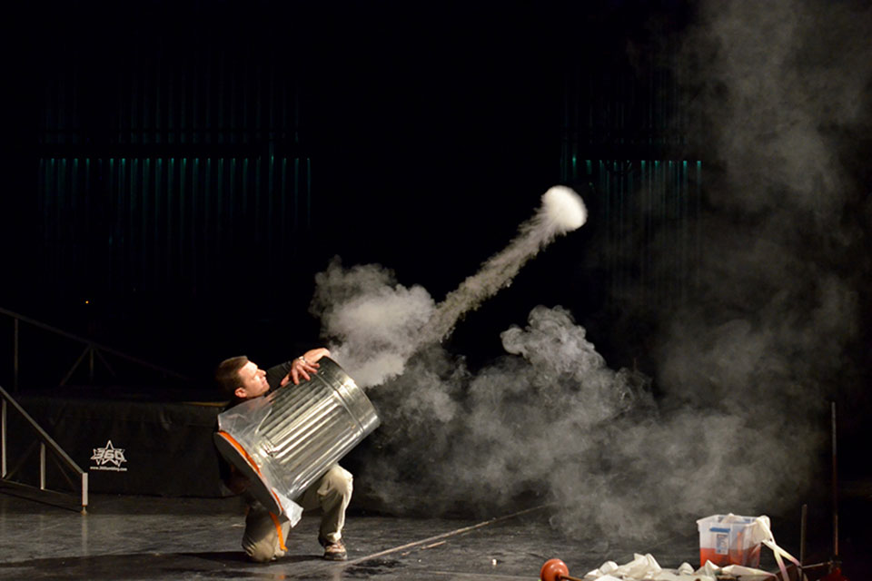 A man on a stage uses a trash can drum to demonstrate physics