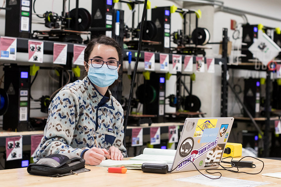 Masked student working at lab table in front of a wall of 3D printers