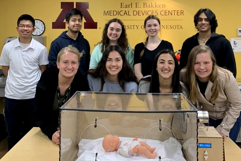 Nine students in front of an incubator with a baby model.