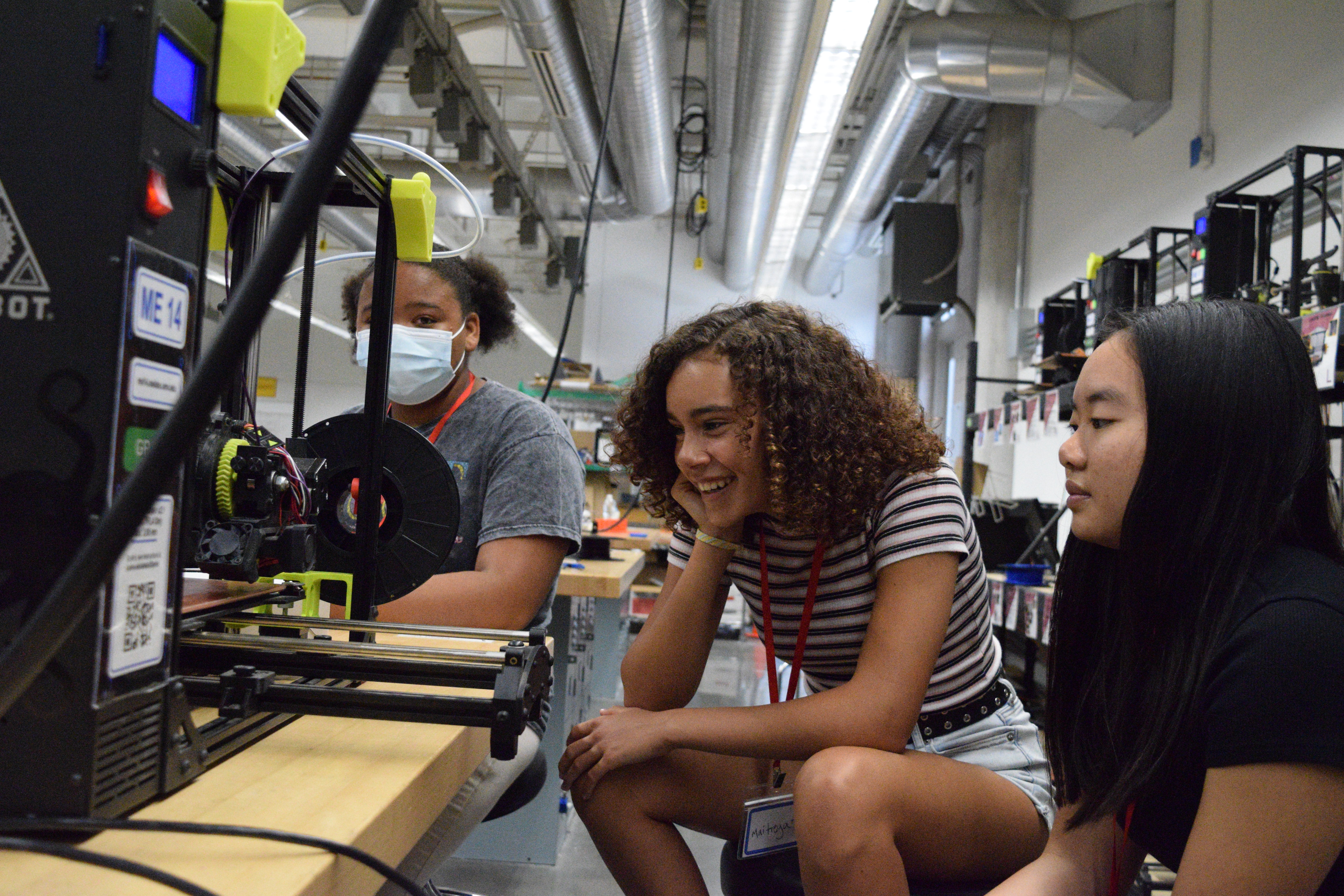 Three young women watching a 3D printer in action.
