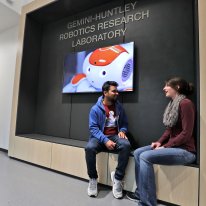 A man and a woman sit talking in front of a monitor with a robot face on it, under a sign that says Gemini-Huntley Robotics Research Laboratory