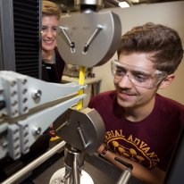 A man in a maroon U of M shirt works on a machine while a woman behind him watches