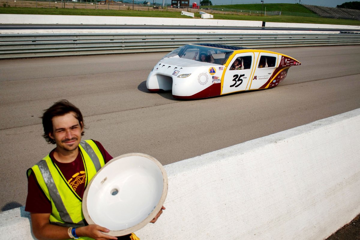 A man wearing a Solar Vehicle Team shirt stands in front of a racetrack where the solar vehicle is racing behind him