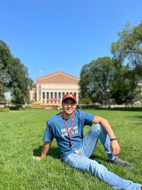 A man wearing a Gophers baseball cap sitting on the lawn of the U of M Mall. Behind him is Northrop Auditorium