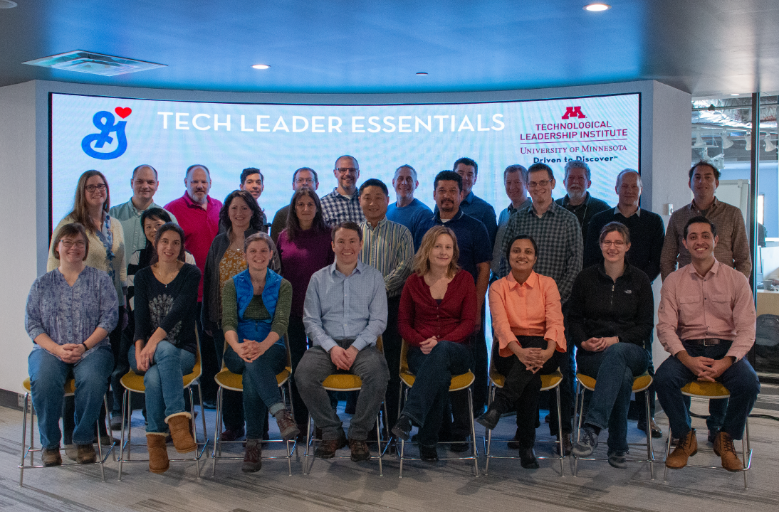 Group of employees from General Mills from a training for their company. There are 26 people split across three rows, the front of which is seated. All are in front of a large digital screen displaying text above them that reads, "Tech Leader Essentials," along with the General Mills and TLI logos.