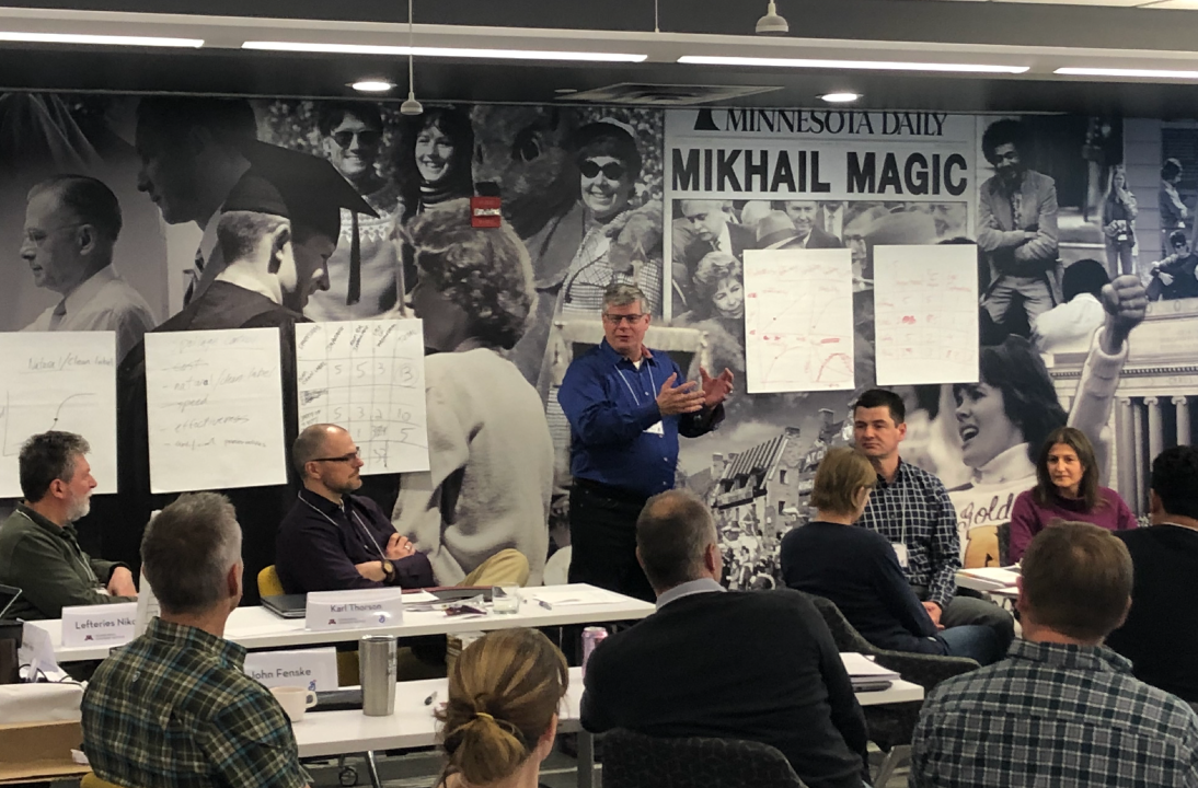 TLI Associate Director Paul Hansen gesturing with his hands as he instructs during a company consulting session. Wall art consists of black and white historical images from events at the U of M, and there are large papers taped up from workgroup brainstorming sessions