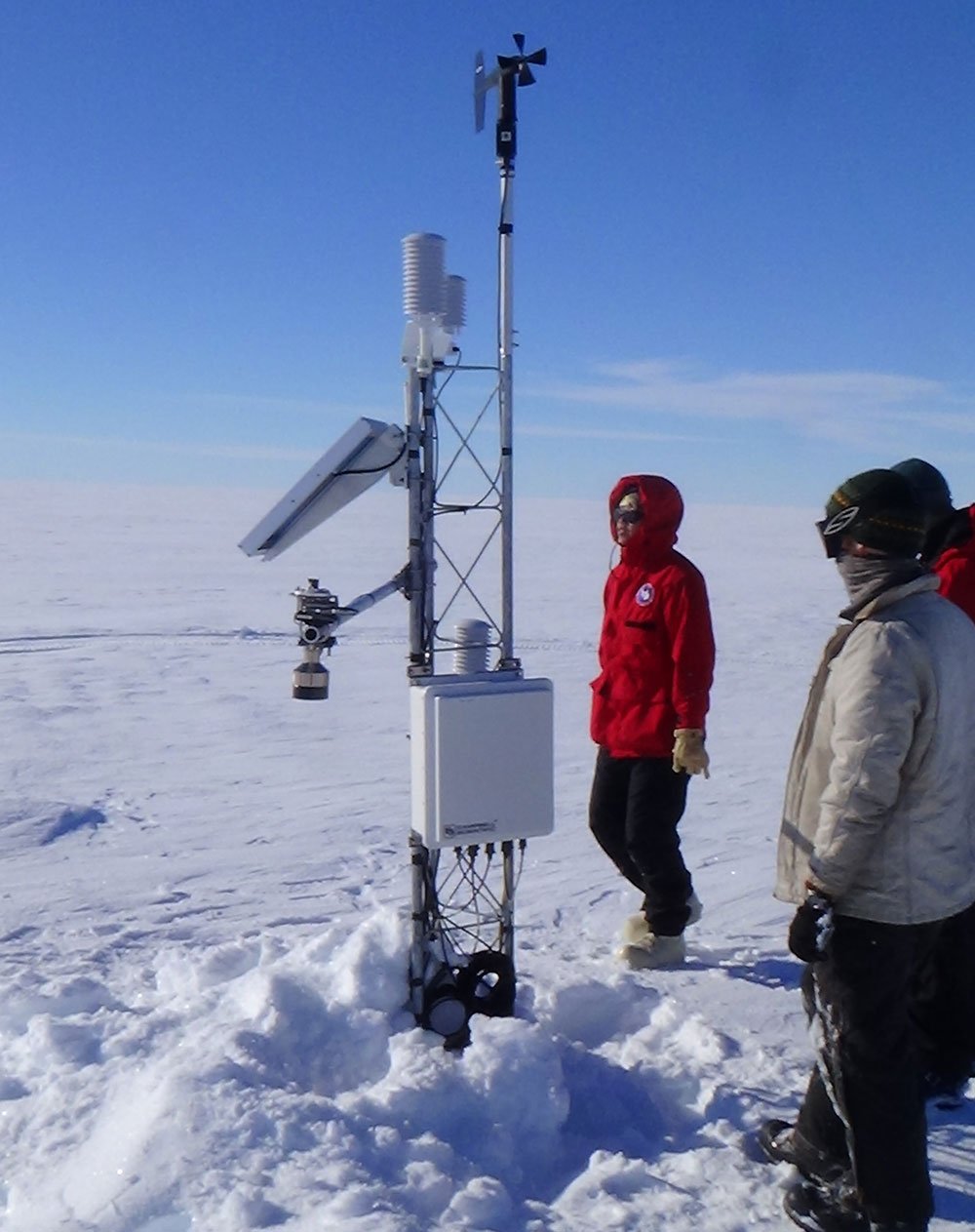 Two people in heavy coats and gear look at a tall sensor array they have just installed in snow