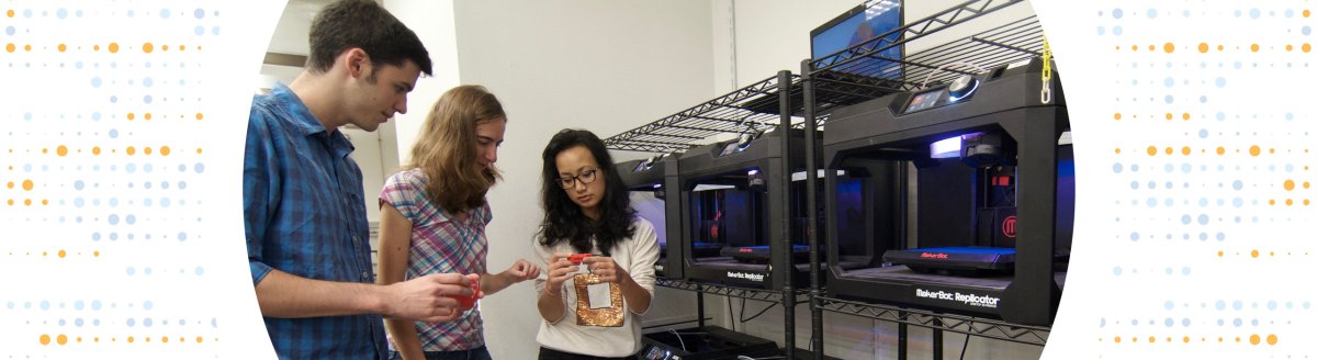 Three students standing in front of a bank of 3D printers, looking at a small red printed block-M