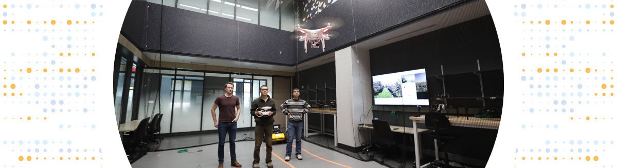Three men piloting a flying drone inside a 2-story lab space