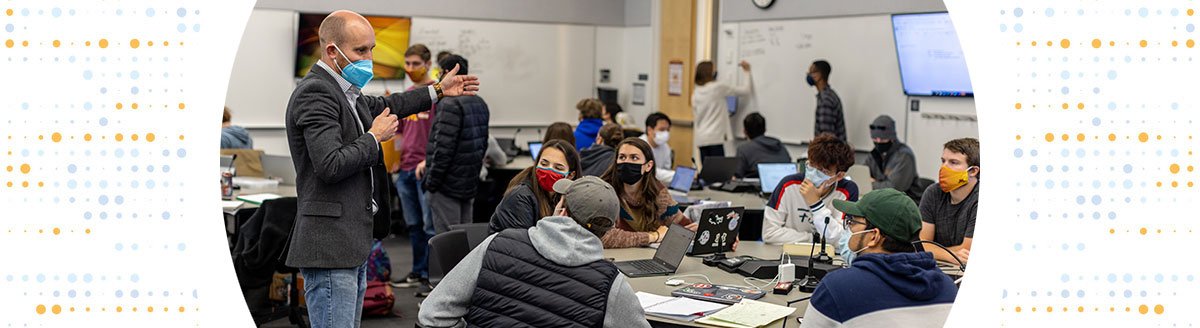A professor instructs a round table full of students, with groups of students using whiteboards and monitors in the background