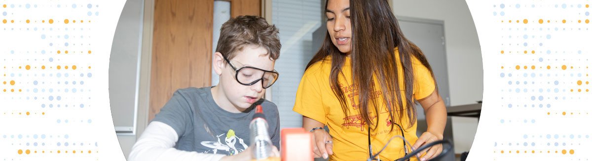 A woman wearing a UM Tech Camp shirt helps a gradeschool student in an engineering lab