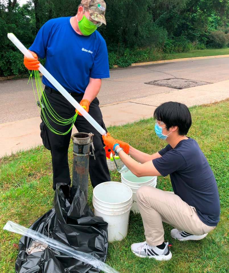 Two men wearing face masks sample fungus outdoors with a long tube and a bucket