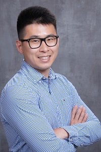 Gang Qiu in a blue gingham shirt standing against a grey background smiling into camera