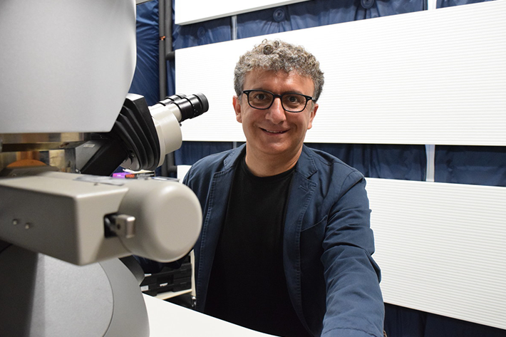 A portrait of Professor Andre Mkhoyan. He wears a blue suit, black glasses, and sits in front of a white microscope. 