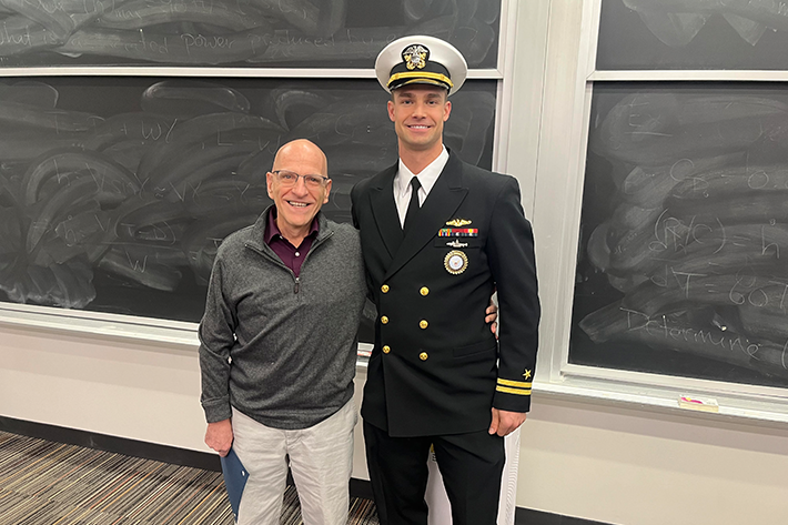 Professor Neurock stands next to the Navy LT who presented him the award in a classroom. The LT is dressed in Navy uniform, and the Professor is dressed in gray.