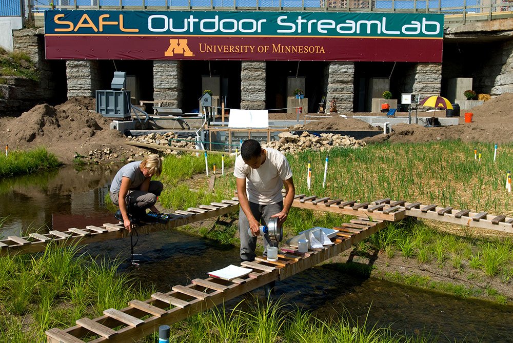 Two students work in grass and water in front of a banner that says Outdoor Stream Lab
