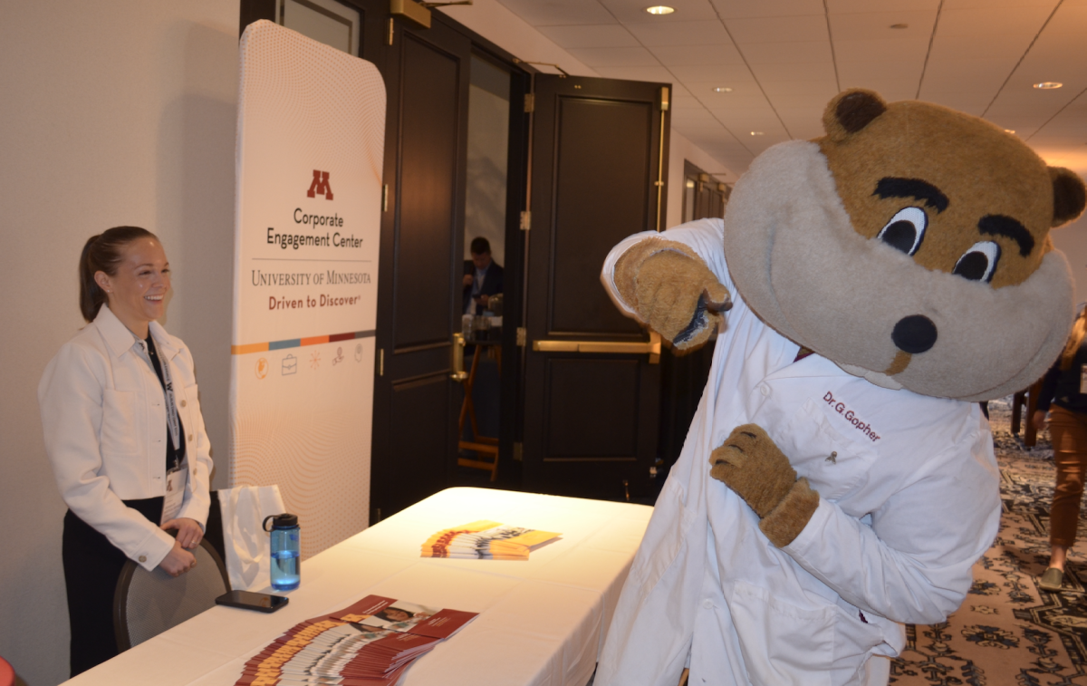 A woman smiles as Goldy Gopher, wearing a lab coat, clowns around at the DMD Conference in Minneapolis