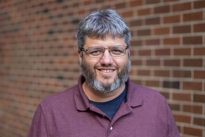A headshot of Prof. Paul D. He wears a maroon polo and stands in front of bricks.