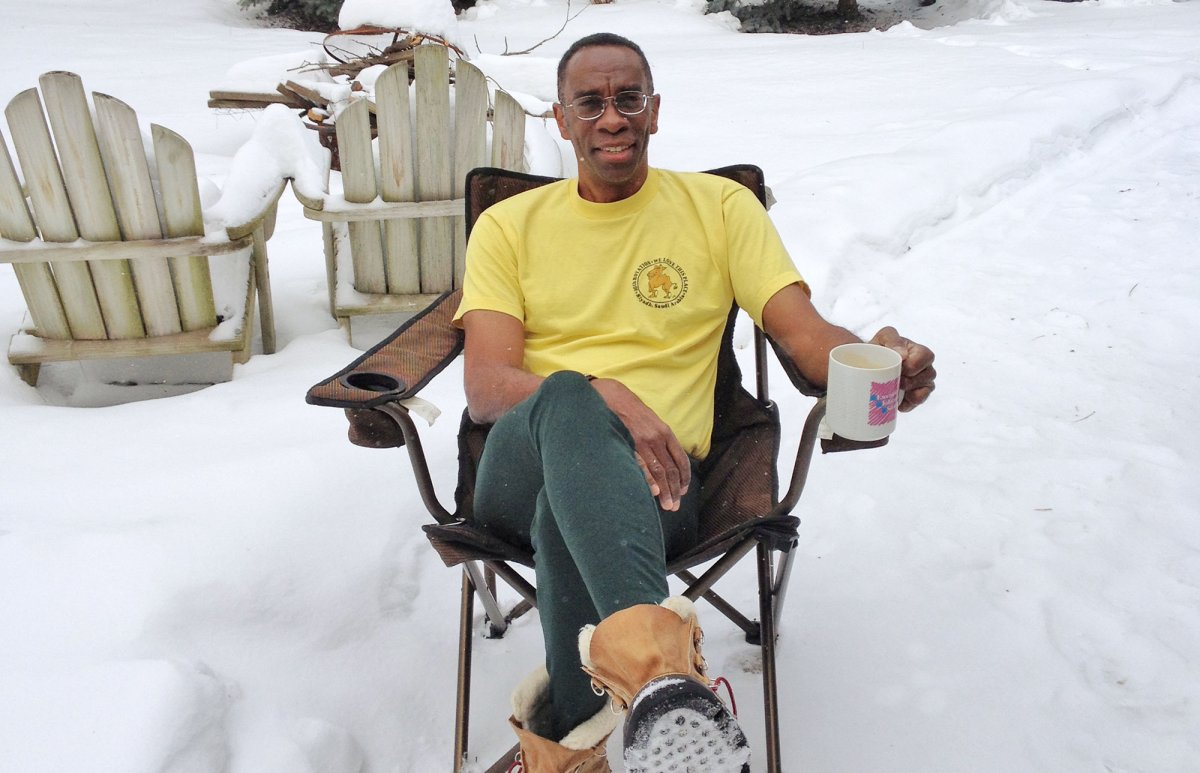 Photo of Adolph Barclift wearing a yellow T-shirt, sitting in a camp chair in the snow, drinking a cup of coffee