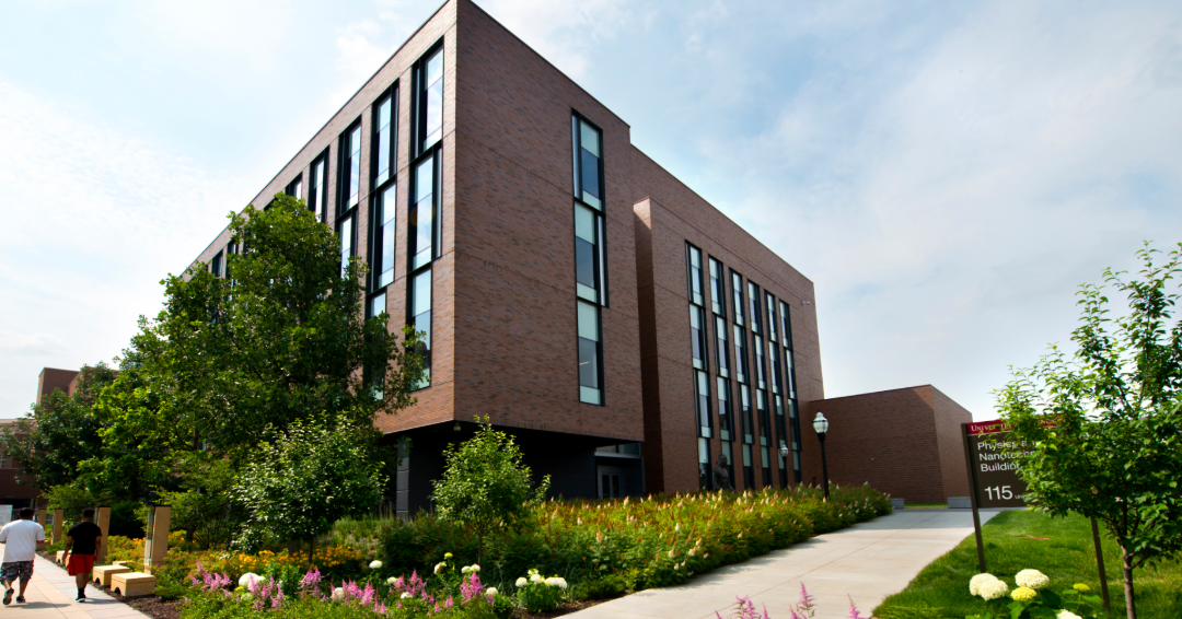 A modern four-story brick building on a college campus with green grass and flowers surrounding it