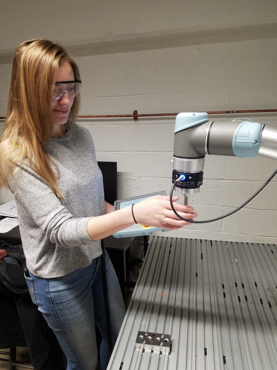 female student in gray shirt with blonde hair and safety goggles handles a robotic arm over a desk