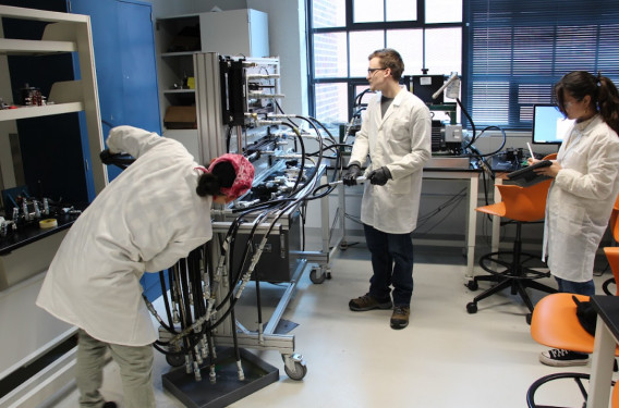 students in lab coats work at a machine with tubes and wires