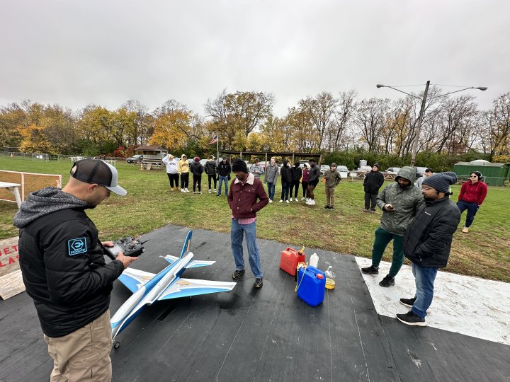 Students stand around a small silver and blue airplane. One student holds a remote control.