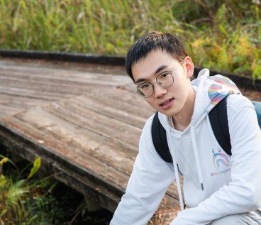 Siliang Zeng outdoors in a white zip up sweatshirt, leaning down smiling into camera