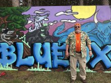 A man in a tye dyed shirt and bandana standing in front of colorful grafitti 