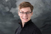 A headshot of student Sage Martin. They wear glasses and a dark colored quarter zip in front of a gray background.