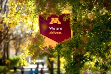 Maroon flag with "We are driven" hanging in a backdrop of trees in autumnal foliage
