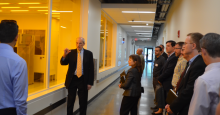 A man in a dark suit, leading a tour, gestures toward a window beyond which is a clean room for tech assembly