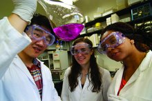 three students in a chemistry lab wearing lab coats and goggle, looking at liquid in a beaker