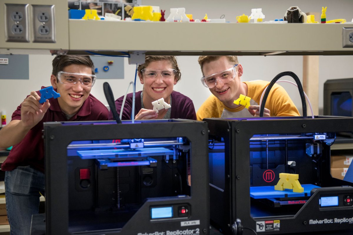 Three students stand behind two 3D printers. They hold up recently printed UMN Ms.