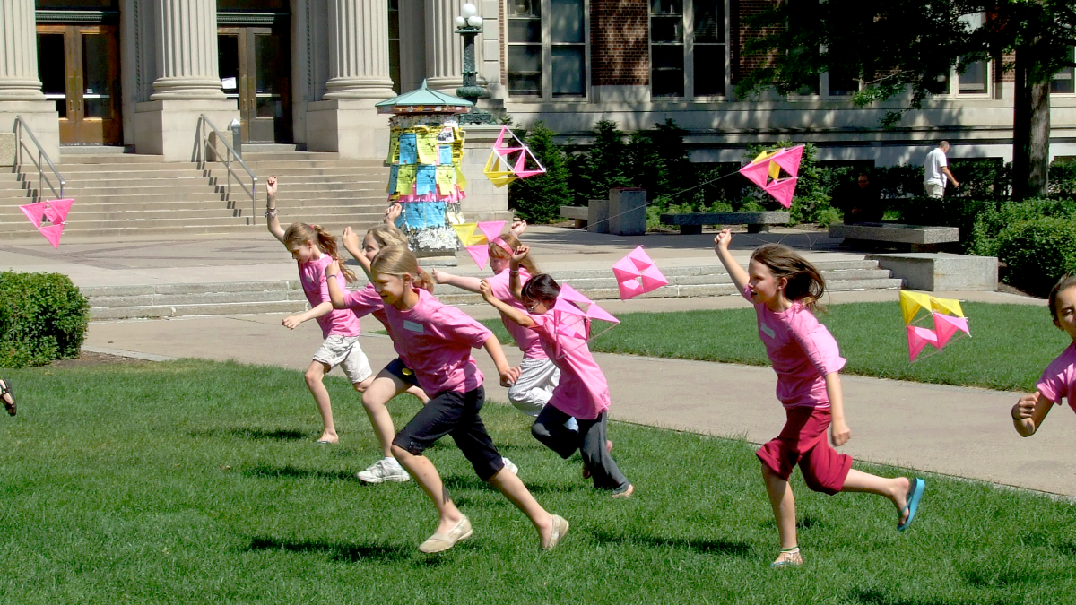 A group of young students in pink shirts run across the campus lawn with geometric kites