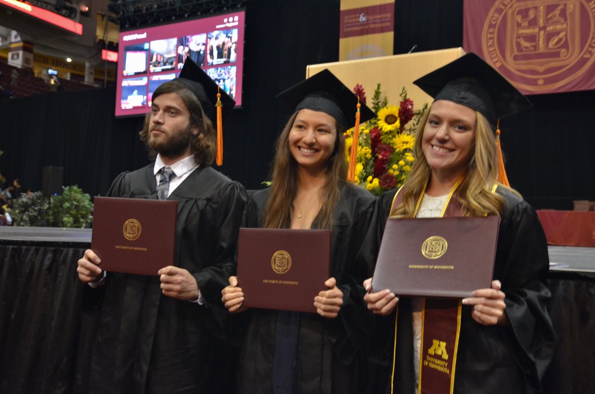 Three students standing in front of stage holding diploma covers