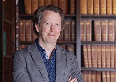 Sean Carroll wearing a blue shirt and blazer standing in front of books