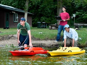Graduate students Josh Vander Hook (l.) and Pratap Tokekar ready two robot boats for launch as Volkan Isler watches from shore
