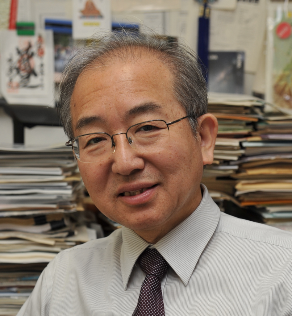 Kazunari Shibata: middle-aged Asian man with glasses and a tie in an office with books and papers behind him