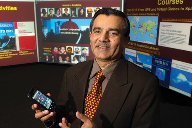 Professor Shashi Shekhar, standing in front of a research poster, holding a smartphone